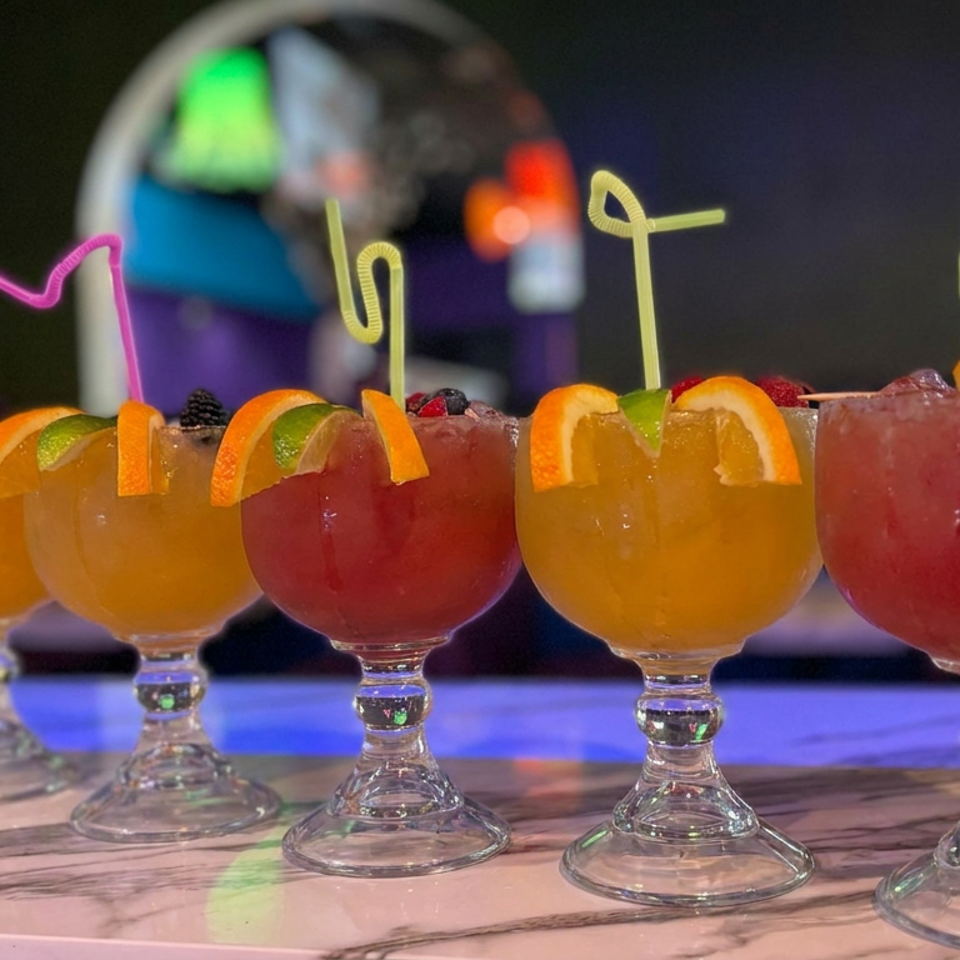 Five colorful cocktails in stemmed glasses garnished with orange and lime wedges, arranged on a bar with neon lighting.