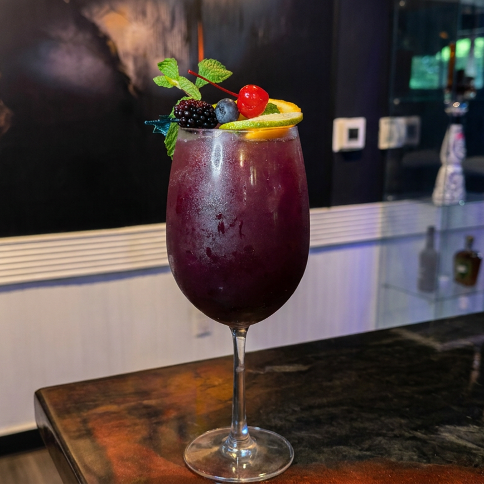 Dark berry cocktail garnished with mint, blackberries, blueberries, cherry, and citrus slice on a polished bar.