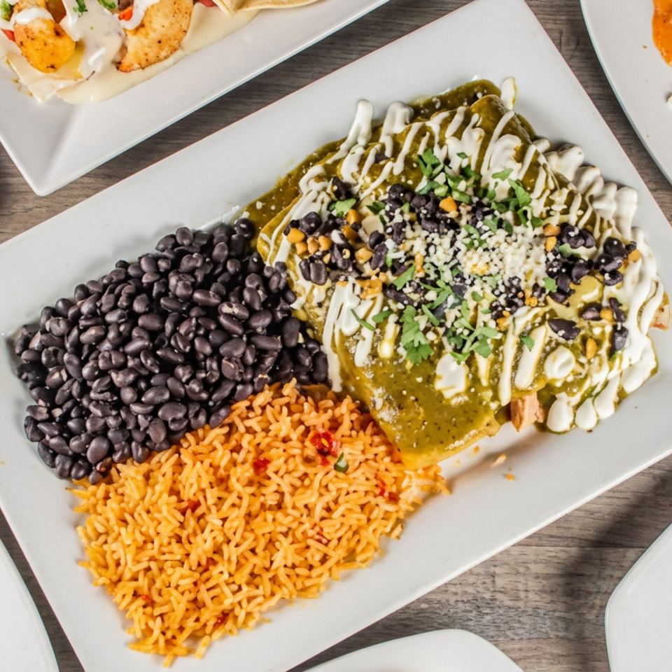 Overhead view of Mexican dishes, featuring enchiladas with green sauce, black beans, and orange rice on a wooden table.